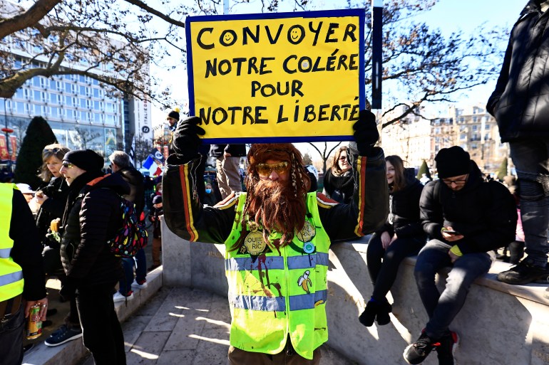 Um manifestante vestindo um colete amarelo segura uma placa que diz "Vamos escoltar nossa raiva por nossa liberdade"