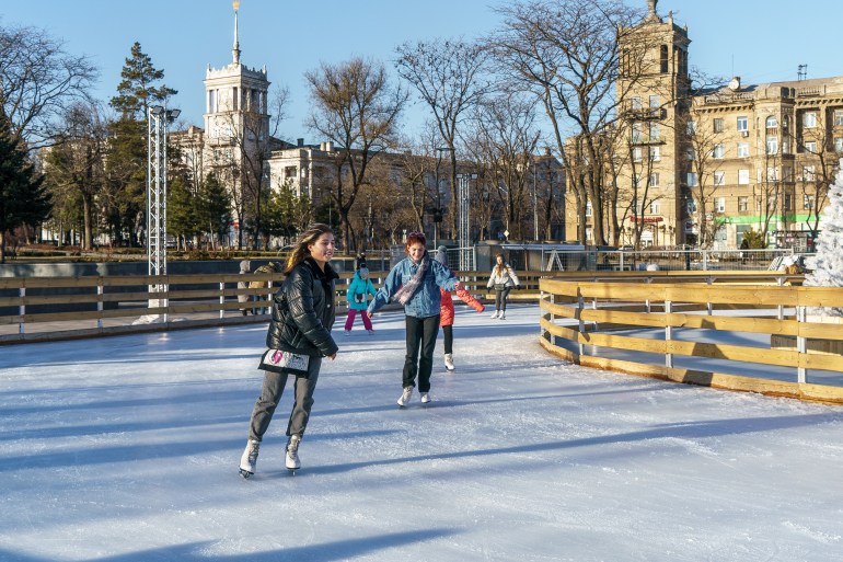 Two teenage girls dressed in warm winter clothes skate on an outdoor rink in the center of Mariupol