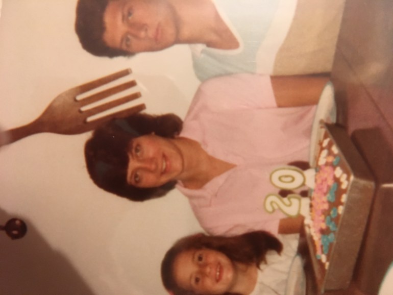 A photo of three people, a woman in the middle with a cake and a candle in the shape of the number 20 in front of her with a man sitting on the right side of the photo and a child sitting on the left.