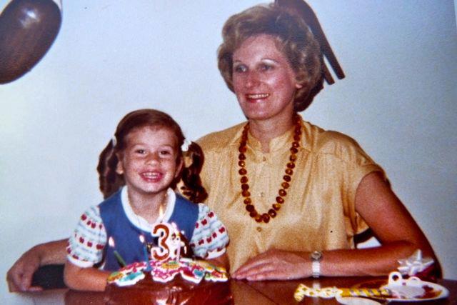 A photo of a child and her mother with a birthday cake in front of the child and a candle in the middle in the shape of the number 3.