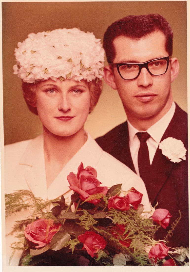 A photo of a woman and a man on their wedding day with the woman wearing a white flowery hat and white outfit and holding a pink rose bouquet and the man wearing glasses and a white flower on his lapel.