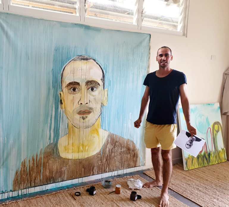 Moz, in black t-shirt and beige shorts - stands in his studio wit a self portrait he has been working on