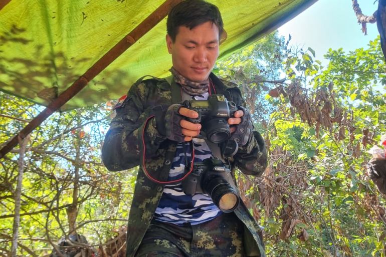 A portrait of Khun Nan Nan standing under a green canopy in the forest, examining his camera.