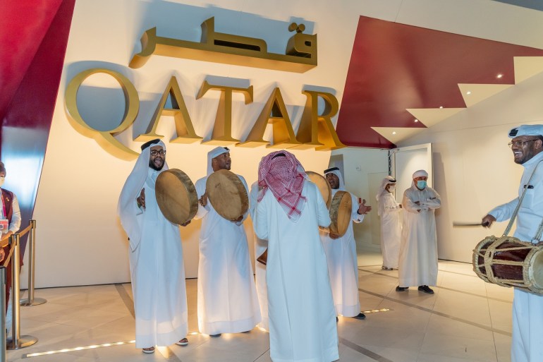 A group of men in traditional dress sing and play the drums celebrating Qatar National Day with 'Qatar' displayed on the wall behind them