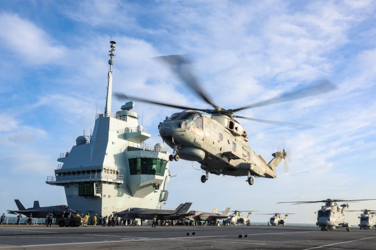 A helicopter takes off from the deck of HMS Queen Elizabeth with the ship's bridge in the background and a blue sky with scattered clouds