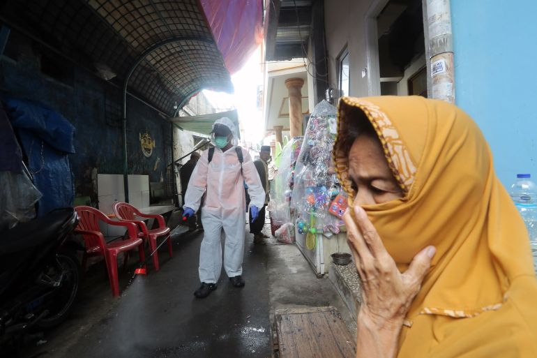 A woman in a yellow headscarf covers her face as a man in a hazmat suit walks along an alley spraying disinfectant