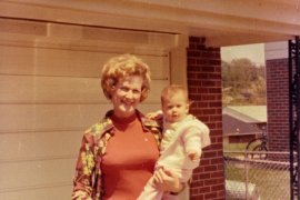 The writer's mother carrying her as a baby, standing in front of a house on a bright sunny day.
