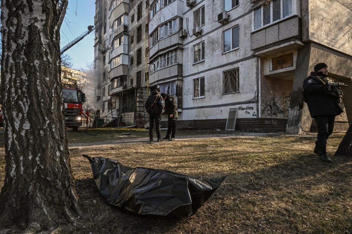 A body in a plastic bag lies on the ground outside a burning building