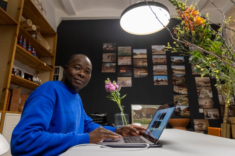 Diebedo Francis Kere, in a royal blue shirt, sits at his desk in his Berlin office