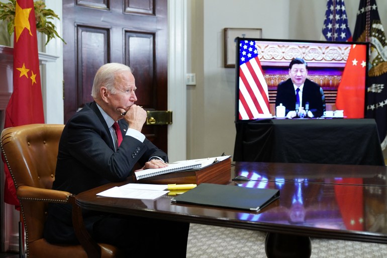 US President Joe Biden sitting at the end of a desk as he holds virtual discussions with Chinese President Xi Jinping, seen smiling on a television screen
