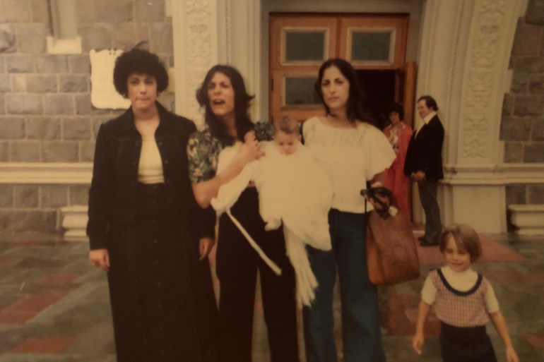 A photo of three women standing outside of a church, the woman in the middle is holding a baby with the child next to them.
