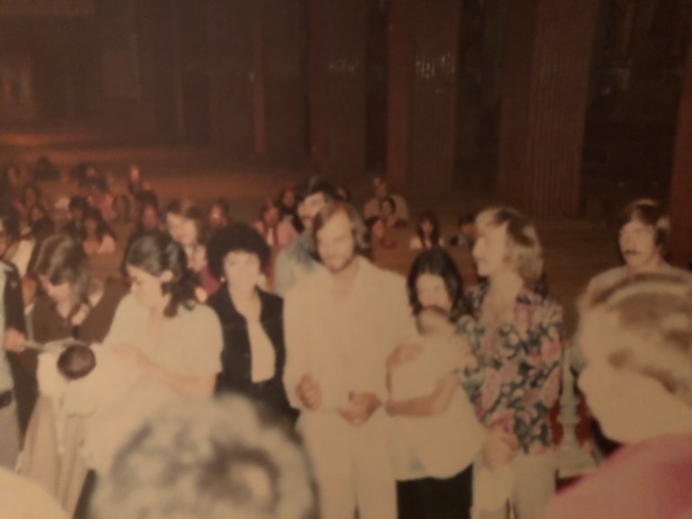 A photo of four people standing in a church full of people. One of them is a woman holding a baby.