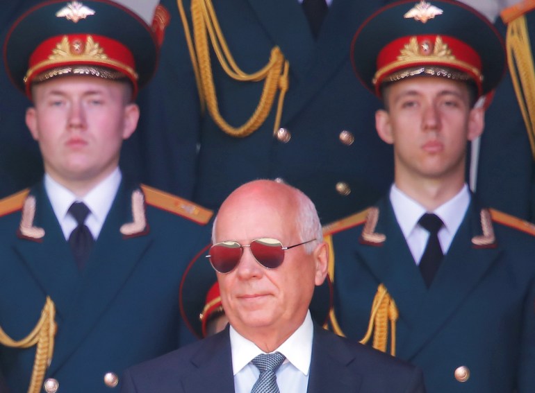 Rostec CEO Sergei Chemezov in mirrored glasses stands in front of two soldiers in uniform