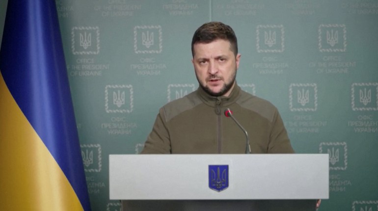 El presidente ucraniano, Volodymyr Zelenskyy, vestido de verde caqui y de pie junto a una bandera ucraniana, pronuncia un discurso en video