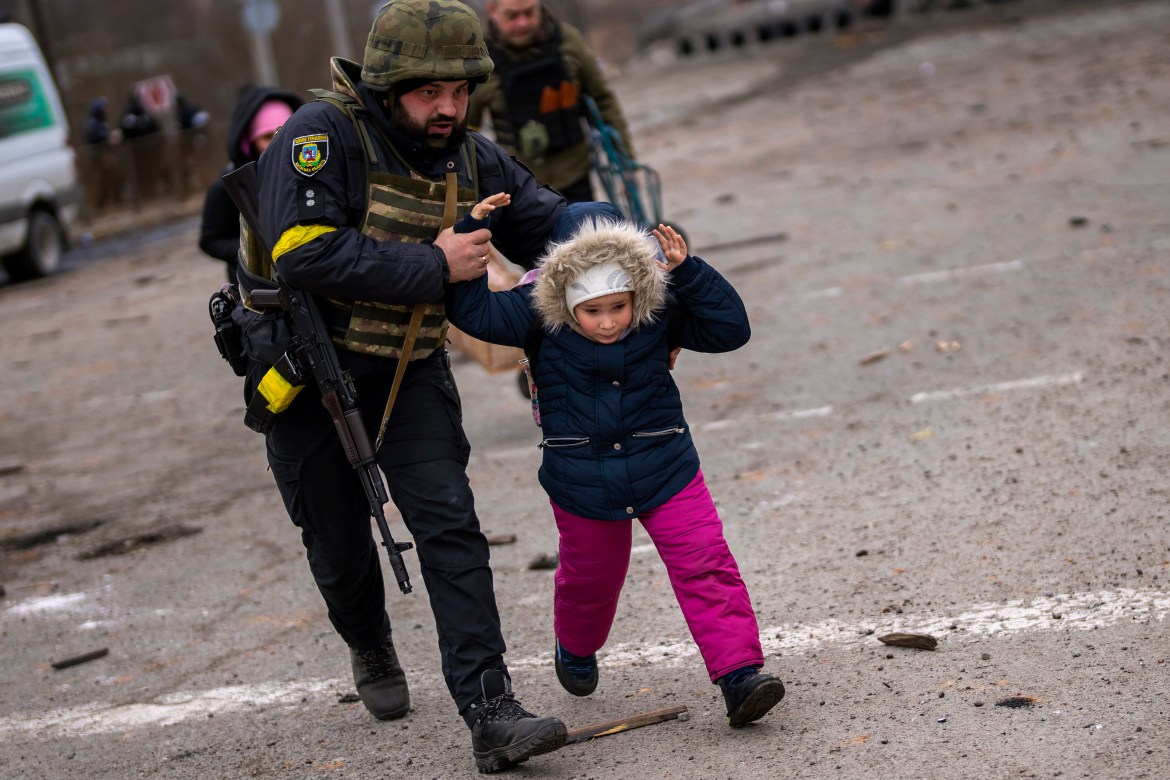 A Ukrainian police officer runs while holding a child as the artillery echoes nearby, while fleeing Irpin on the outskirts of Kyiv