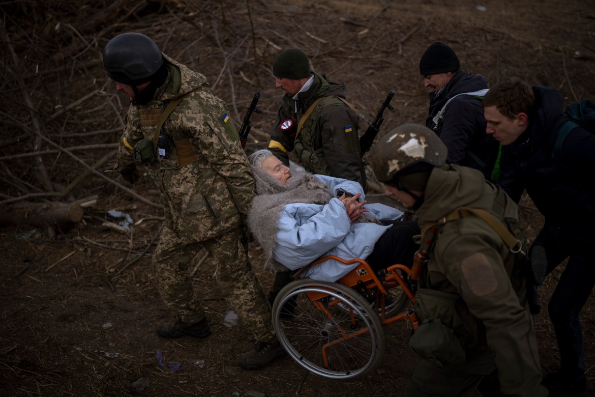 Ukrainian soldiers and militiamen carry a woman in a wheelchair as the artillery echoes nearby, while people flee Irpin on the outskirts of Kyiv