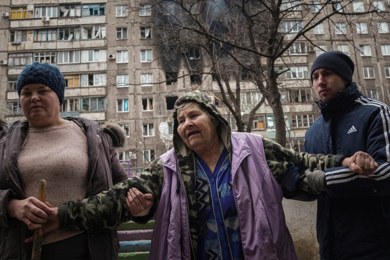 People help an elderly woman to walk in a street with an apartment building hit by shelling in the background in Mariupol