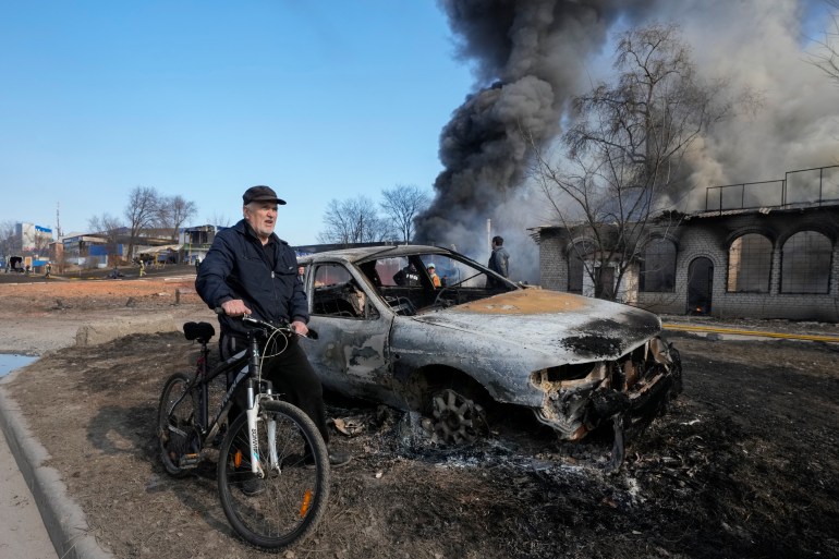 A man rides his bike past the gutted remains of a car and smoke rising from a fire following a Russian attack in Kharkiv
