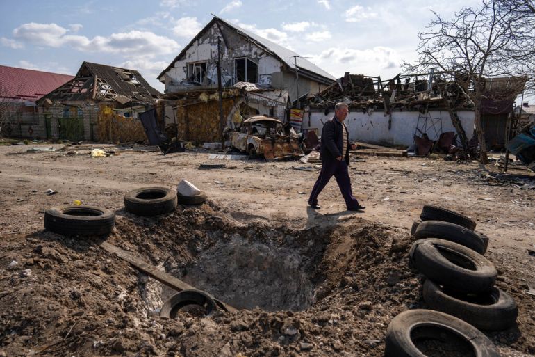 A man walks behind a crater created by a bomb and in front of damaged houses.