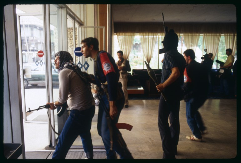 Several armed men stand in the lobby of a hotel looking out through the glass doors and windows to the street beyond