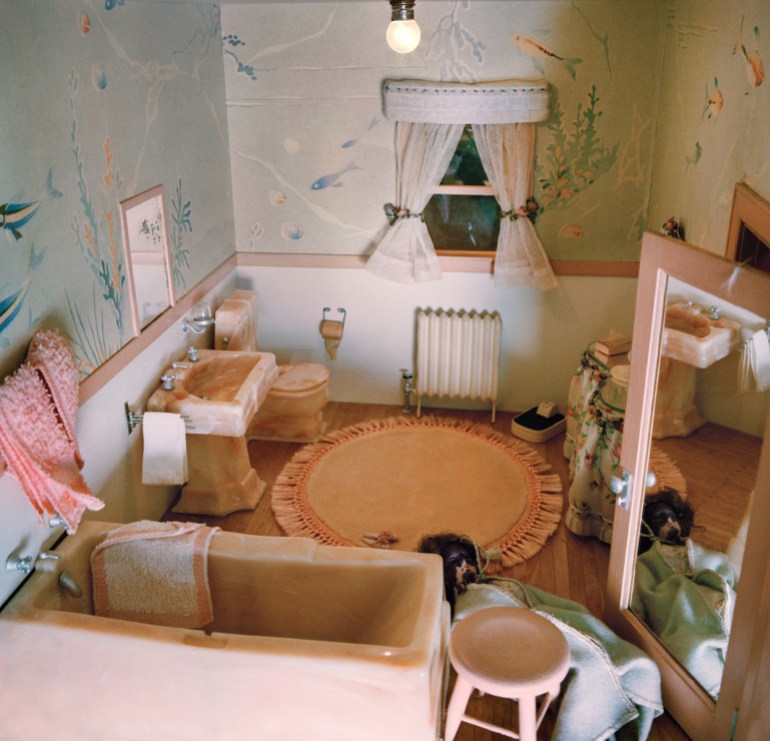 A photo of a diorama of a bathroom with a doll lying on the floor by the bathtub in front of a mirror behind the bathroom door.