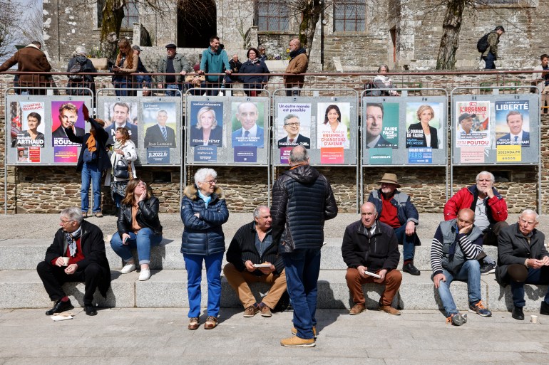 Local residents stand on the main square, next to the twelve official posters of presidential candidates.