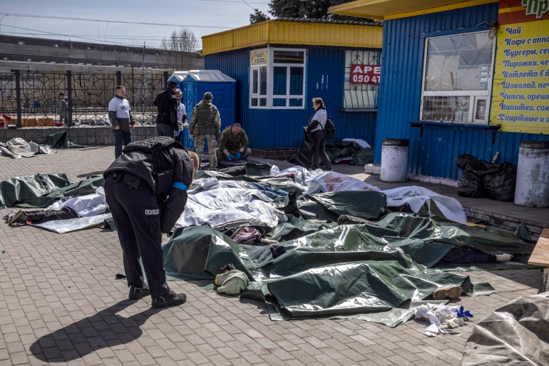 A Ukrainian policeman bends over bodies laid on the ground and covered with tarpaulin