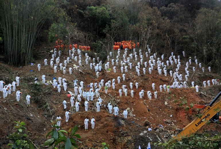 Recovery teams in white suits dotted around a muddy hillside against a forest as they pay a silent tribute to those who died on the China Eastern plane that crashed in Wuzhou, Guangxi Zhuang Autonomous Region, China, in March 2022.
