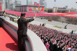 Kim Jong Un in black suit waves from behind a balcony to tens of thousands of people gathered in formation in Pyongyang's Kim Il Sung Square.