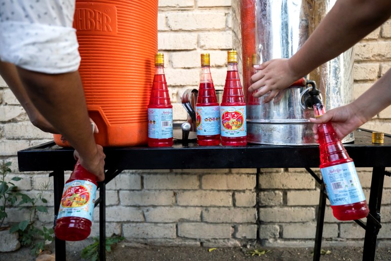 Two large dispensers of prepared rooh afza are seen on a table with two sets of hands filling bottled with the drink.