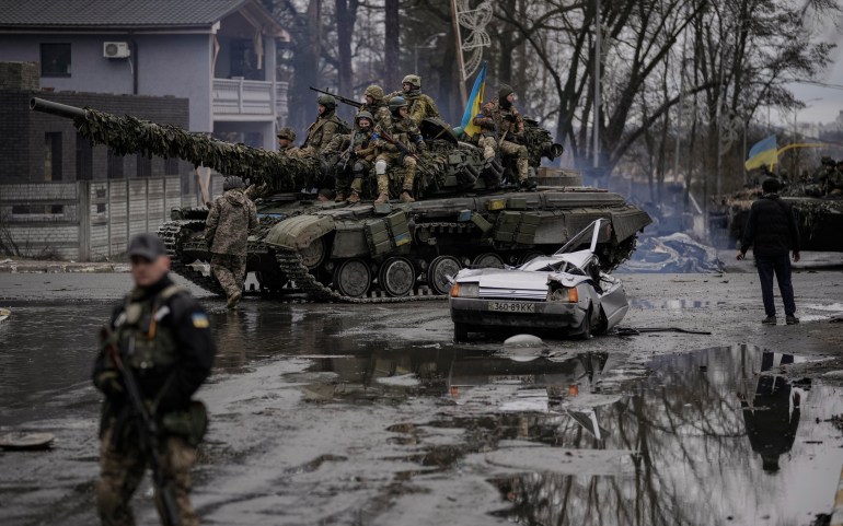 A man stands next to a civilian vehicle destroyed in the fighting between Ukrainian and Russian forces with the driver's body still dead. 
