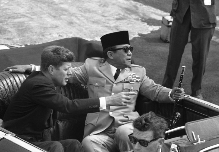 Sukarno lounges across on the back seat of an open top car next to John F Kennedy on a visit to the US