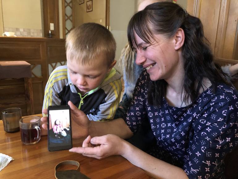 A photo of a woman holding a phone with a photo of Olena is holding a lamb and a child is behind the phone leaning to look at what is on the screen.