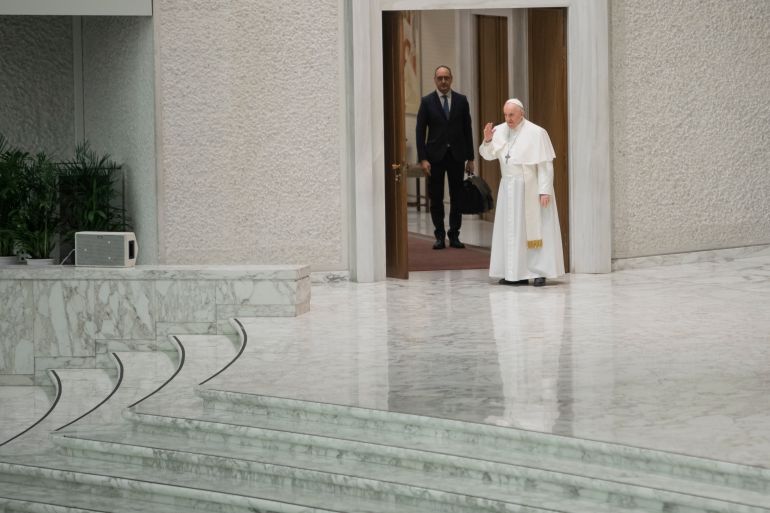 A photo of Pope Francis waving with a man in a suit standing behind him.