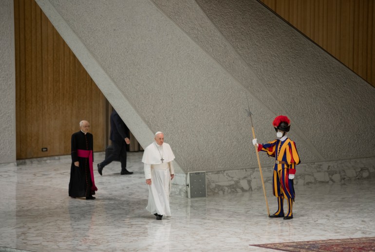 A photo of Pope Francis walking with a bishop behind him and a security guard standing in front of him.
