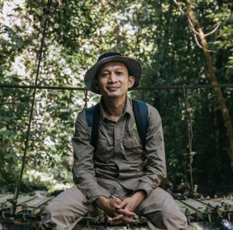 Rudi Putra, wearing a khaki green rangers uniform and hat, sits surrounded by forest