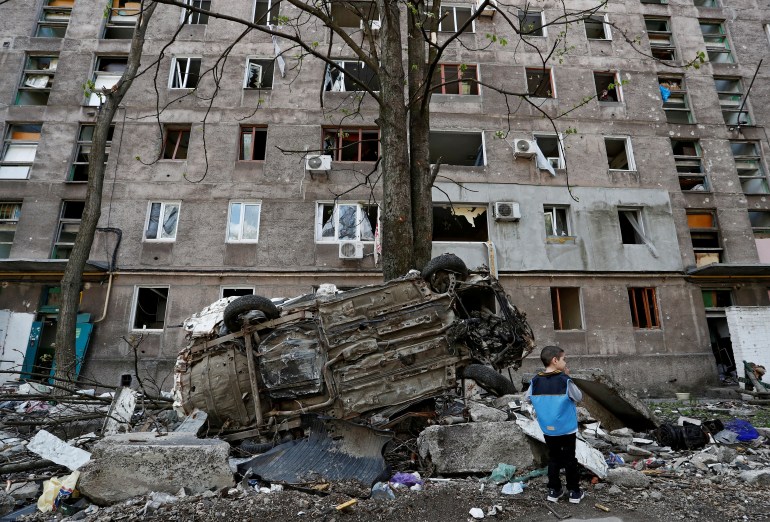 A boy stands next to wrecked vehicle in front of an apartment building damaged during Ukraine-Russia conflict in the southern port city of Mariupol.