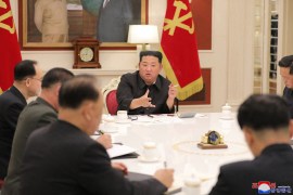Kim Jong Un in Mao-style suit in front of party flags berates officials sat with him around a table.