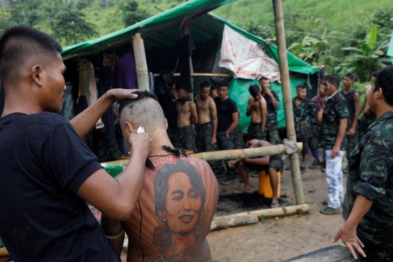 A member of the PDF, with his back to the camera showing a massive tattoo of Aung San Suu Kyi, gets his head shaved at a KNU training camp in the jungle as other recruits watch from a bamboo hut with a roof of green tarpaulin