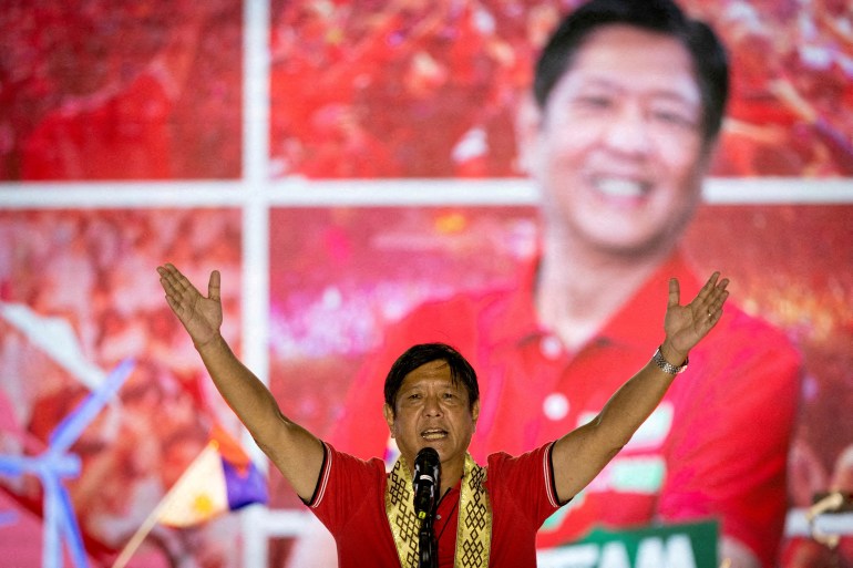 Philippine presidential candidate Ferdinand "Bongbong" Marcos Jr in a reo shirt with a garland around his neck holds his arms aloft to acknowledge the crowd at a rally 