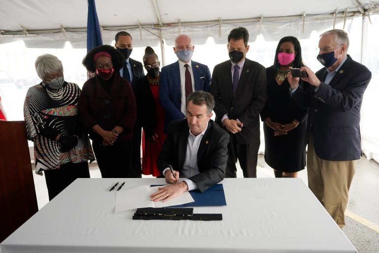 Virginia Gov. Ralph Northam seated at a table and with activists and legislators standing behind him signs the law abolishing the death penalty