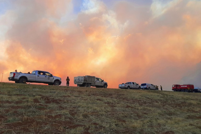 Fire is seen in the background behind several cars and vans