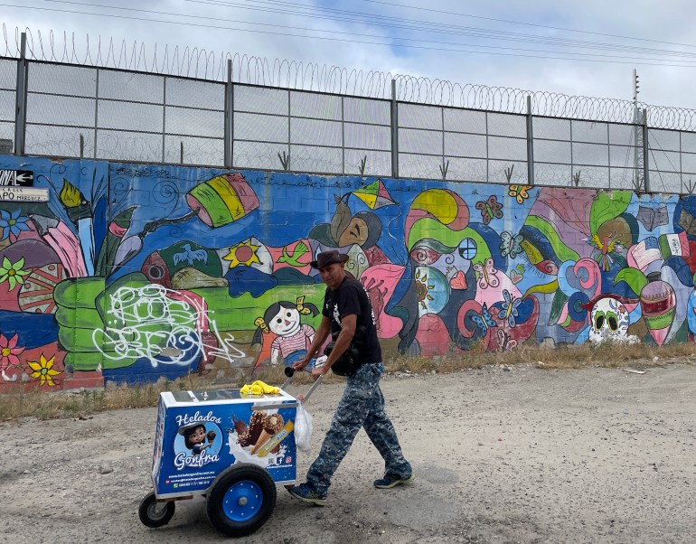 A man walks with an ice cream cart next to the border wall in Tijuana that separates Mexico from the US.