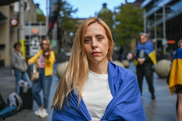 Ukrainian refugee Olesia, who has long blonde hair and is wearing a white t-shirth with her country's blue and yellow flag around her shoulders, at a rally for Ukraine in Sydney's Martin Place