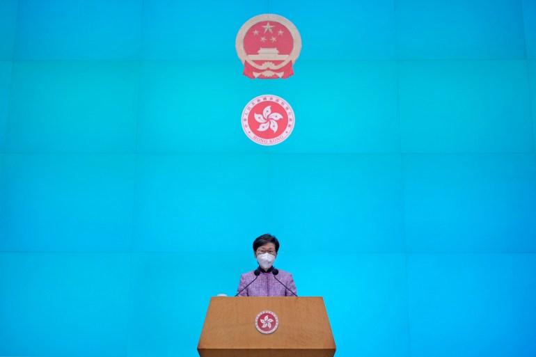 Carrle Lam se tient à une conférence devant un mur bleu vif avec les armoiries de Hong Kong et le symbole du Parti communiste chinois derrière elle
