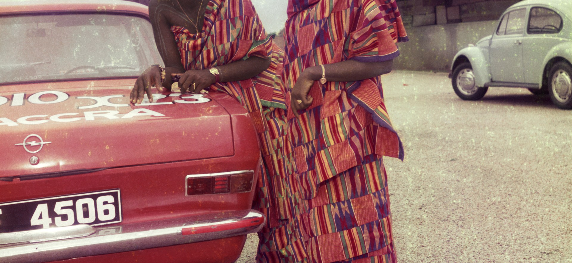 Two women dressed for a church celebration stand next to a red car