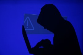 A hooded man holds a laptop computer as blue screen with an exclamation mark is projected on him
