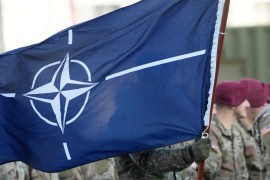 NATO flag with troops in formation in the background.