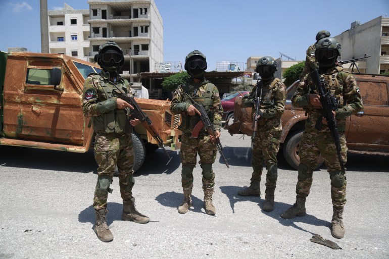 Four fighters in camouflage uniforms and face masks on stand with their guns in front of vehicles