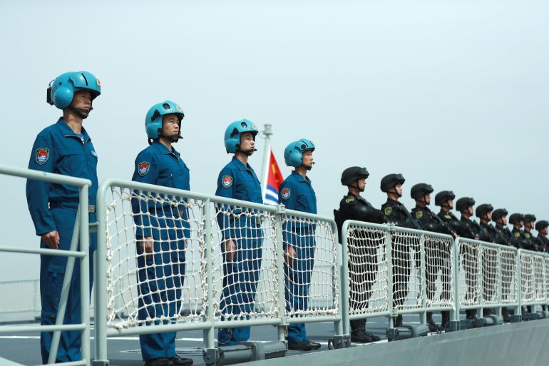 A photo of members of the Chines Navy stand on the deck of a navy ship at a military port.